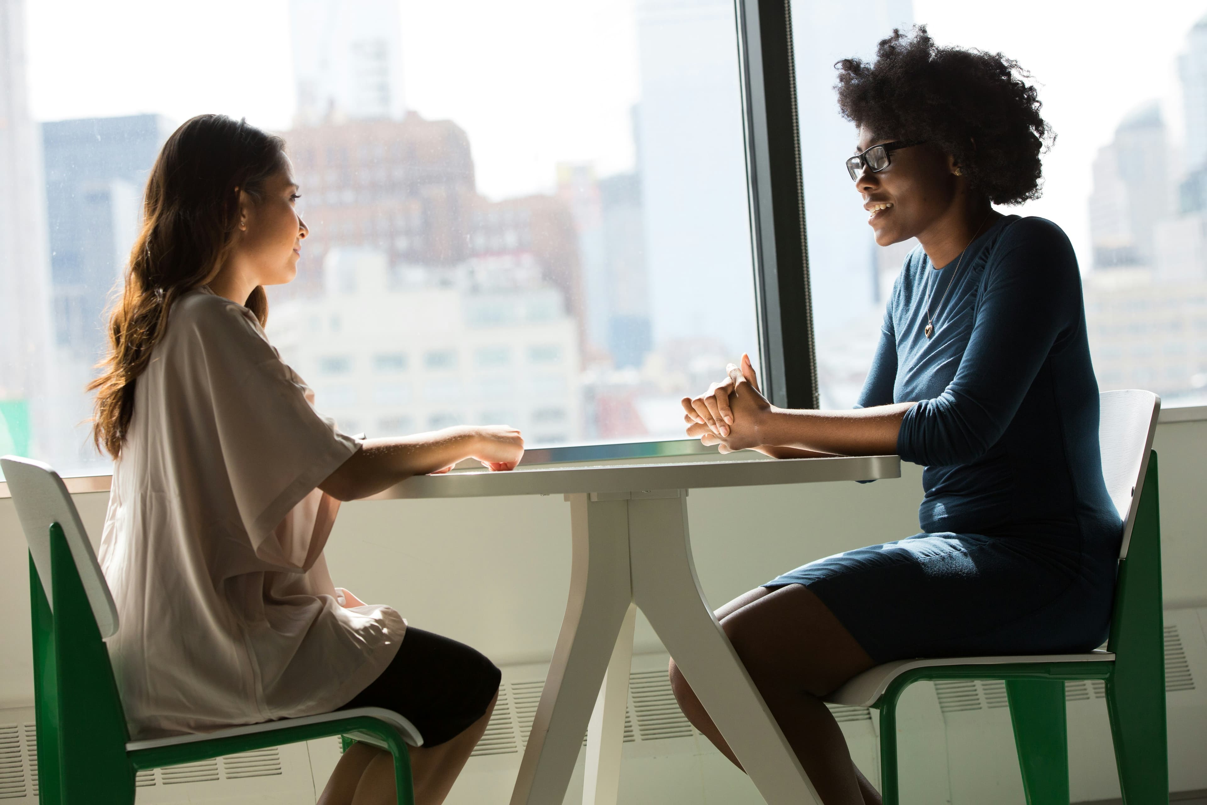 Two businesswomen in conversation at a table beside a window overlooking a city skyline